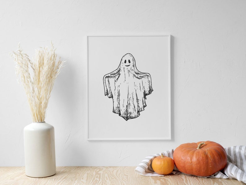 May include: A black and white illustration of a ghost with a smiling face in a white frame. The frame is on a white wall with a vase of dried pampas grass and two pumpkins on a wooden table.