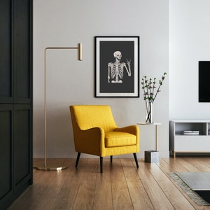 May include: A mustard yellow upholstered armchair with black legs positioned next to a tall gold floor lamp and a small side table holding a glass vase with green branches. A framed wall art print depicts a white skeleton on a dark gray background.