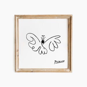 May include: A framed print with a black line drawing of a butterfly on a white background. The artwork is signed "Picasso" in black script. The frame is a light brown colour.
