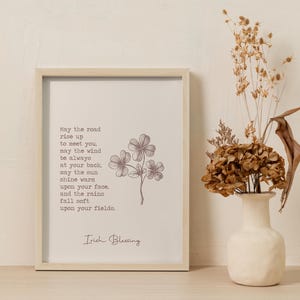 May include: A framed print with an Irish blessing, featuring a floral illustration. The text reads "May the road rise up to meet you..." in a serif font. A vase with dried flowers sits to the right.