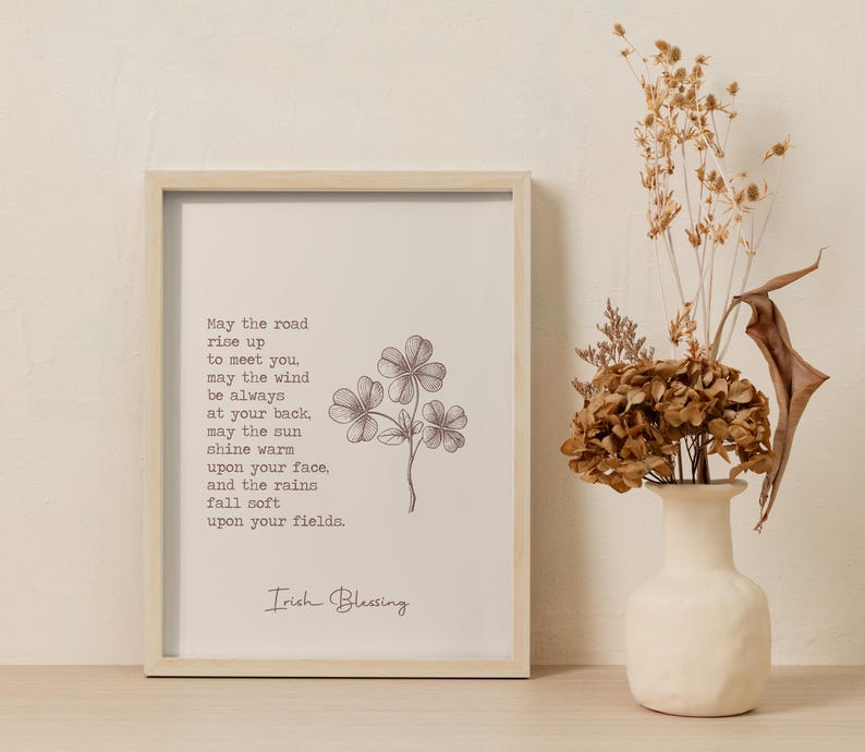 May include: A framed print with an Irish blessing, featuring a floral illustration. The text reads "May the road rise up to meet you..." in a serif font. A vase with dried flowers sits to the right.