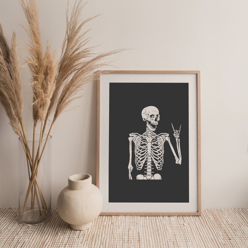 May include: A framed wall art print featuring a white skeleton illustration on a dark gray background. The skeleton is depicted from the waist up, with its mouth open and one hand raised in a rock and roll hand gesture with fingers extended.