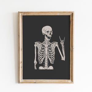 May include: A framed wall art print featuring a light beige skeleton illustration against a solid dark charcoal background. The skeleton is depicted from the chest up, with its mouth open and one hand raised in a rock and roll hand gesture.