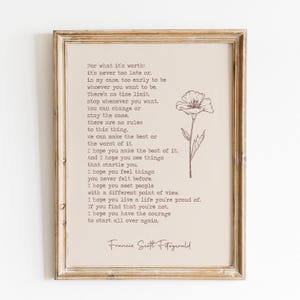 May include: A framed wall art print on a light beige background featuring the text of a quote by F. Scott Fitzgerald in a typewriter-style font, accompanied by a simple line drawing of a single flower on the right side, all in a wooden frame.