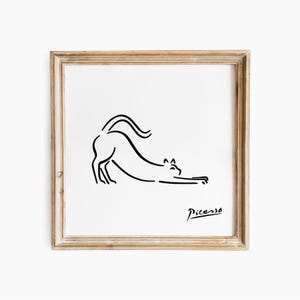 May include: A framed print with a minimalist black line drawing of a stretching cat. The artwork is signed "Picasso" in the lower right corner. The frame is light brown, and the background is white.