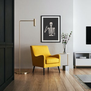 May include: A yellow armchair with black legs sits in a living room with a black and white framed print of a skeleton making a peace sign. A gold floor lamp and a small side table with a vase of flowers are also visible.