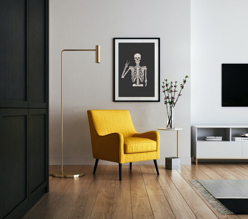 May include: A yellow armchair with black legs sits in a living room with a black and white framed print of a skeleton making a peace sign. A gold floor lamp and a small side table with a vase of flowers are also visible.