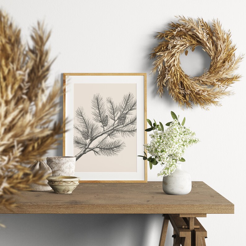 May include: A framed wall art print featuring a black ink sketch of a pine branch with cones on a light beige background. The print is displayed alongside a dried grass wreath, a small white vase with white flowers, and two small ceramic bowls.