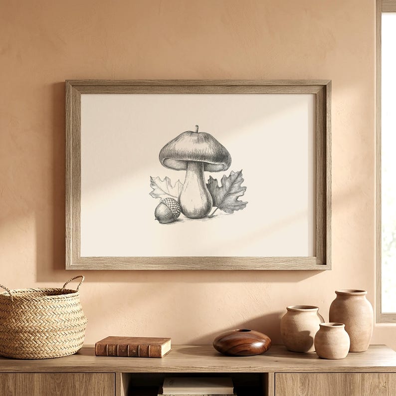 Pode incluir: Uma pe&ccedil;a de arte de parede emoldurada apresentando um desenho a l&aacute;pis de grafite de um cogumelo, uma bolota e duas folhas de carvalho centralizados num fundo claro. A obra est&aacute; inserida numa moldura retangular de madeira com acabamento natural.