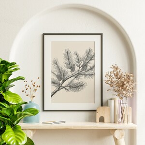 May include: A framed wall art print featuring a black and white line drawing of a pine branch with three pinecones on a solid beige background. The artwork is enclosed in a thin black rectangular frame, presenting a botanical illustration style.