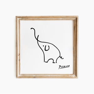 May include: A framed art print with a minimalist black line drawing of an elephant on a white background. The artwork is signed "Picasso" in the lower right corner. The frame is a light brown colour.
