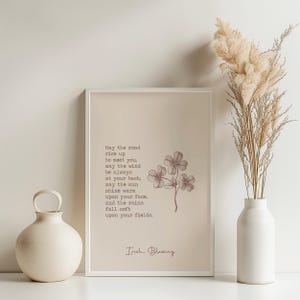 May include: A framed print with an Irish blessing, featuring a four-leaf clover illustration. The text reads "May the road rise up to meet you..." A beige vase and dried ornamental grass are on the right, and a round vase with a handle is on the left.