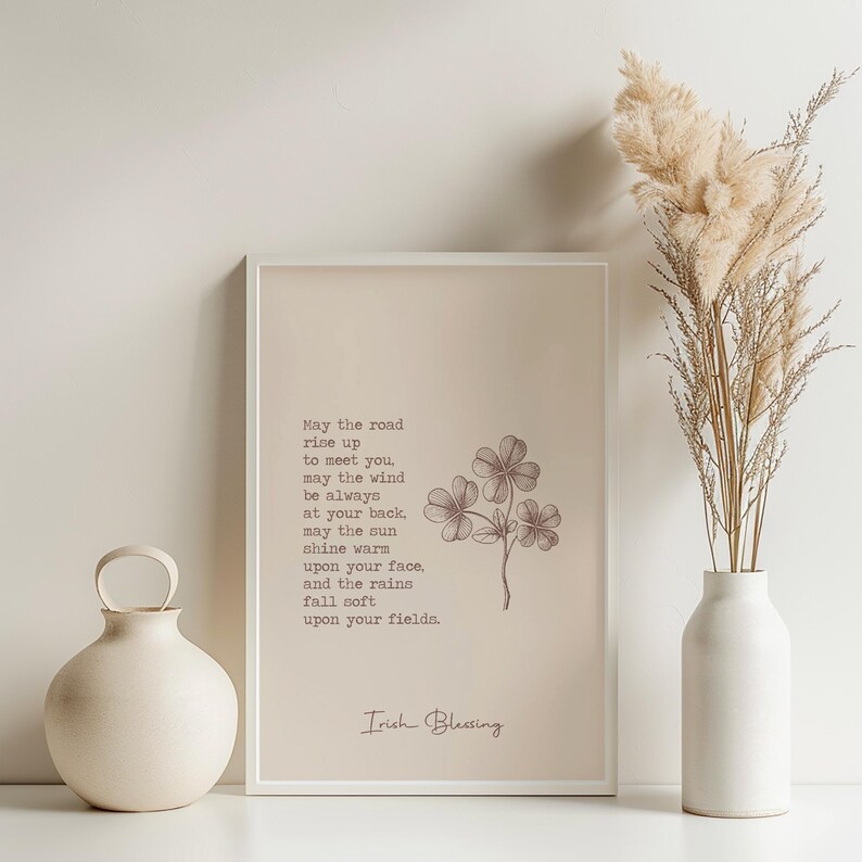 May include: A framed print with an Irish blessing, featuring a four-leaf clover illustration. The text reads "May the road rise up to meet you..." A beige vase and dried ornamental grass are on the right, and a round vase with a handle is on the left.