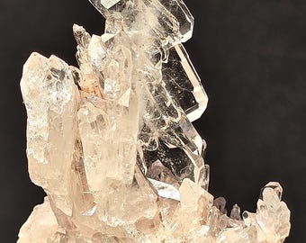 May include: A cluster of clear quartz crystals with a pink hue, arranged in a tower-like formation. The crystals are set on a base of rough, gray rock.