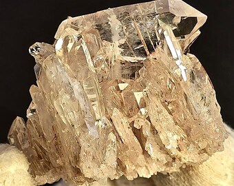 May include: A cluster of clear quartz crystals with a light brown hue. The crystals are arranged in a cluster, with some of the crystals pointing upwards and others pointing downwards. The crystals are translucent and have a glassy luster.