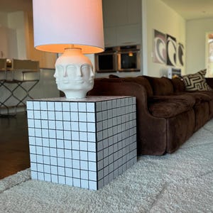 May include: A white lamp with a white lampshade sits on a white and black tiled cube end table. The lamp features a white sculpture of three faces. The table is in front of a brown leather sofa.