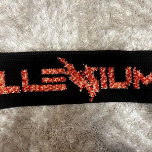 May include: A rectangular black knitted panel featuring the word ILLENIUM in a textured, variegated red, orange, and white yarn. The letters are stitched into the black background, with a lightning bolt symbol replacing the letter N.