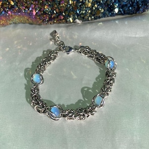 May include: A silver-toned chainmail bracelet with six light blue opalite beads. The bracelet is made of interwoven metal rings and has a lobster clasp closure. The opalite beads are set in wire cages.