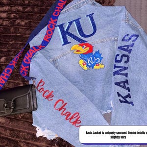 May include: Light blue denim jacket with distressed details. Features the letters "KU" and "KANSAS" in blue glitter, along with a cartoon Jayhawk mascot. The words "Rock Chalk" are in red glitter on the sleeve.