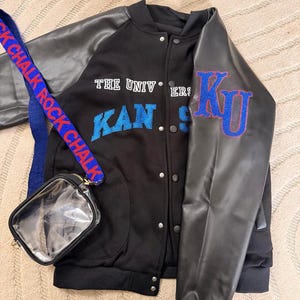 May include: Black varsity jacket with gray faux leather sleeves. The jacket features the text "THE UNIV" and "KANSAS" in blue, with a "KU" patch on the sleeve. A small clear purse with a blue and red strap that says "ROCK CHALK" is also in the image.