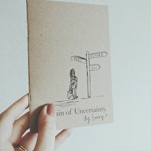 May include: A brown paper notebook with a hand-drawn illustration of a person standing at a crossroads with signs pointing to "Past", "Future", and "Joy". The notebook cover reads "Chain of Uncertainty. By Emsy."