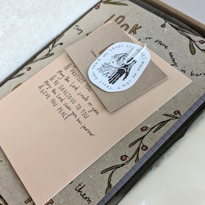 May include: A pink card with the text "& Protect you. May the Lord smile on you & be gracious to you. May the Lord show you his favour & give you peace."  A white sticker with a hand drawn illustration of two hands holding each other and the text "Hand we trust his hair. When we can't" and the website address "www.tracegods.com".