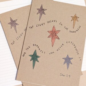 May include: Two brown notebooks with a quote from John 1:5, "The light shines in the darkness, and the darkness can never extinguish it." The quote is surrounded by five stars in different colors.