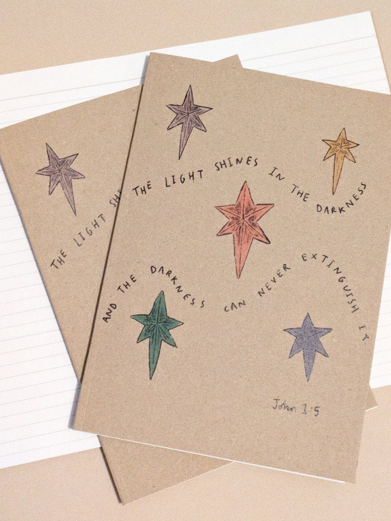 May include: Two brown notebooks with a quote from John 1:5, "The light shines in the darkness, and the darkness can never extinguish it." The quote is surrounded by five stars in different colors.