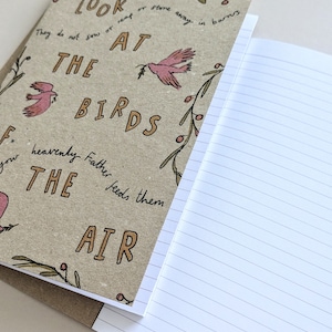 May include: A brown notebook with a quote about birds and a floral design. The quote reads "Look at the birds, they do not sow or reap or store away in barns, your heavenly Father feeds them, the air."