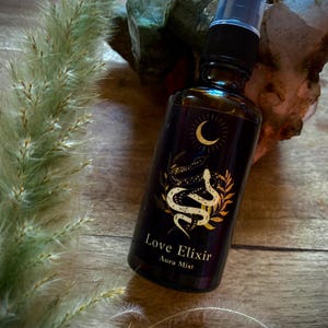Puede incluir: Una botella de vidrio marrón oscuro con un pulverizador negro, etiquetada como "Love Elixir Aura Mist". La etiqueta presenta una luna creciente dorada y un diseño de serpiente. La botella está rodeada de plantas secas y cristales.