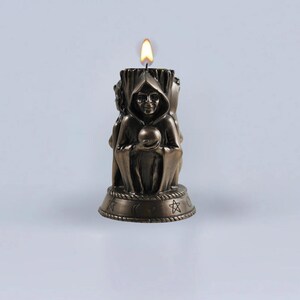 May include: A bronze candle with a three-headed figure holding a sphere. The candle is lit and has a base with symbols including a crescent moon, a star, and a circle.