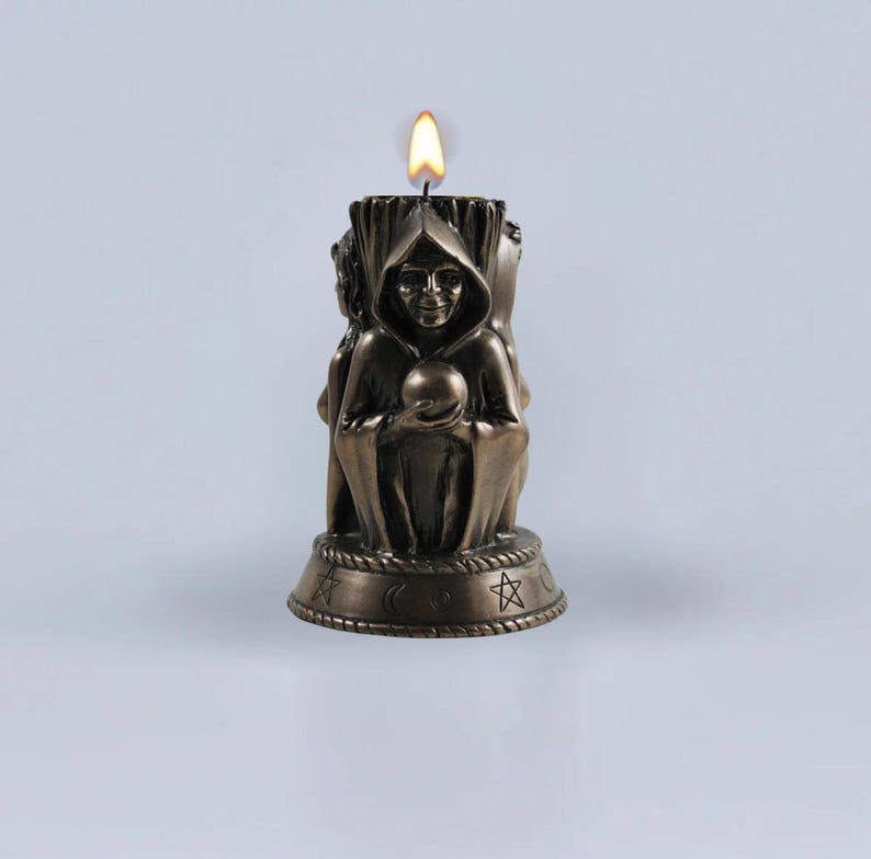 May include: A bronze candle with a three-headed figure holding a sphere. The candle is lit and has a base with symbols including a crescent moon, a star, and a circle.