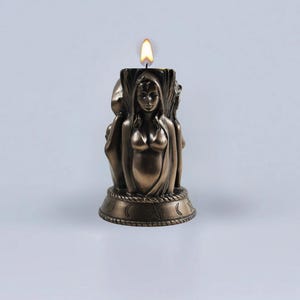 May include: A bronze candle holder featuring a three-faced goddess figure with a lit candle in the center. The base of the holder is decorated with moon phases and a pentagram.