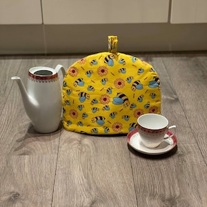 Könnte beinhalten: Ein gelber Tee-Wärmer mit einem Bienen- und Sonnenblumenmuster. Der Wärmer steht auf einem Holzboden mit einer weißen Teekanne und einer weißen Teetasse mit Untertasse.