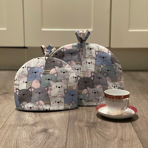 May include: Two grey and blue tea cosy covers with a pattern of cartoon cats. The covers are on a wooden floor with a white teacup and saucer.