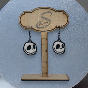 May include: A pair of black dangle earrings with white embroidered Jack Skellington faces from the Nightmare Before Christmas movie. The earrings are hanging from a wooden earring display stand.