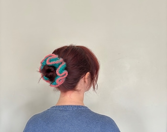 May include: A person with long, dark red hair wearing a blue sweater. Their hair is pulled back in a bun and secured with a teal and pink crocheted hair scrunchie.