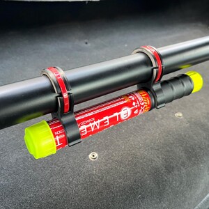 May include: A black cylindrical object with red and black accents is secured to a black pole with red and black clamps. A red and white cylinder with yellow ends is attached to the black pole. The text "E L E M E" is visible on the red cylinder.