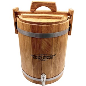Könnte beinhalten: Ein holzfassförmiger Getränkespender mit Zapfhahn. Der Spender hat einen Deckel und einen Griff. Das Fass besteht aus hellbraunem Holz und hat Metallbänder. Der Text "Dancing Rainbow River Cottage" ist auf der Vorderseite aufgedruckt.