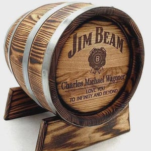 May include: A wooden barrel with metal bands and a wooden stand. The front of the barrel features the "Jim Beam" logo and the text "Charles Michael Wagoner LOVE YOU TO INFINITY AND BEYOND". The barrel is made of light and dark brown wood.