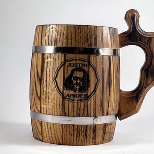 May include: A wooden beer stein with a metal band around the top and bottom. The stein has a handle and is engraved with the words "Justin Abides" and a cartoon image of a man with long hair and sunglasses.