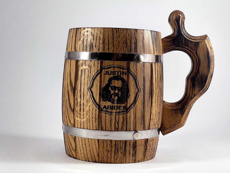 May include: A wooden beer stein with a metal band around the top and bottom. The stein has a handle and is engraved with the words "Justin Abides" and a cartoon image of a man with long hair and sunglasses.