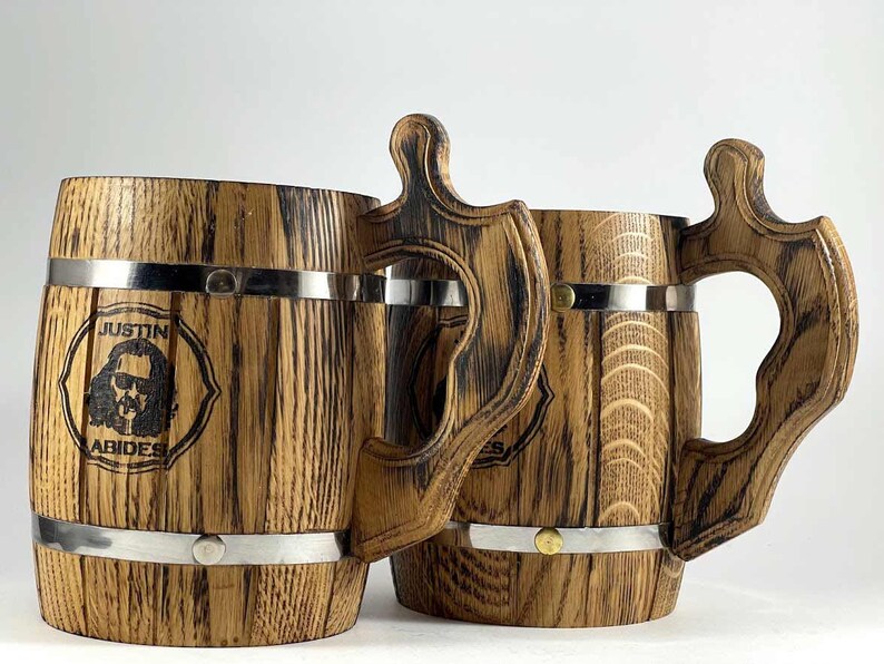 May include: Two wooden beer steins with metal bands and handles. The stein on the left has a personalized design with the text "Justin Abides".