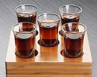 May include: A wooden shot glass holder with six clear shot glasses filled with a dark amber liquid. The shot glasses are arranged in a circular pattern, and the holder is made of light brown wood.