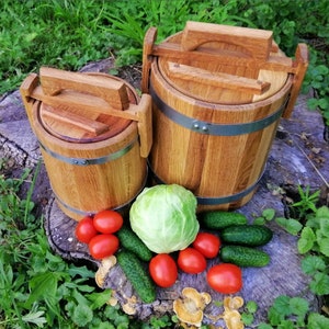 May include: Two wooden barrels with lids and handles, one larger than the other, sitting on a tree stump with green grass and a cabbage, cucumbers, and tomatoes around them.