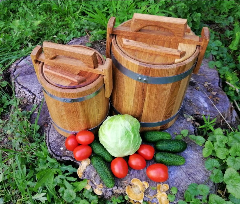 May include: Two wooden barrels with lids and handles, one larger than the other, sitting on a tree stump with green grass and a cabbage, cucumbers, and tomatoes around them.