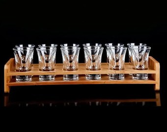 May include: A set of eight clear shot glasses with a geometric design, displayed on a two-tiered bamboo serving tray. The shot glasses are evenly spaced and ready for use. The background is black.