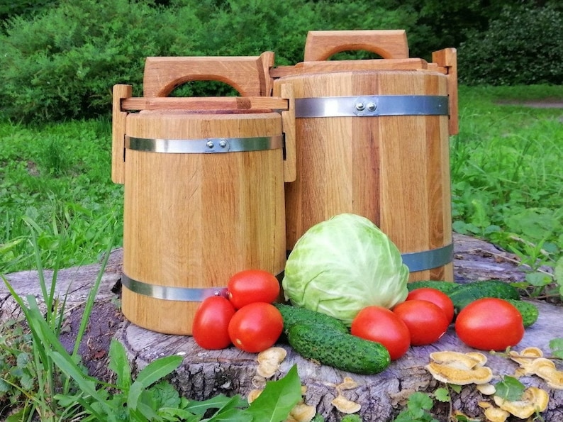 May include: Two wooden barrels with metal bands and handles, one larger than the other, sitting on a tree stump with a cabbage, cucumbers, and tomatoes around them.