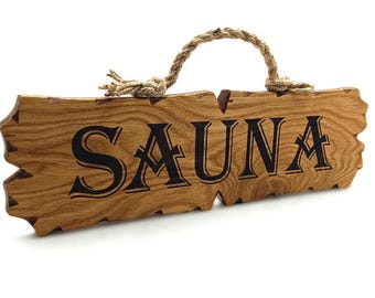 Puede incluir: Un letrero de madera con un asa de cuerda que dice "SAUNA" en letras negras. El letrero está hecho de madera marrón claro con un acabado rústico.