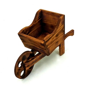 Può includere: Una piccola carriola in legno con finitura rustica in legno bruciato. La carriola ha una singola ruota, una vasca profonda e un manico. La fioriera è progettata per piccole piante o oggetti decorativi.
